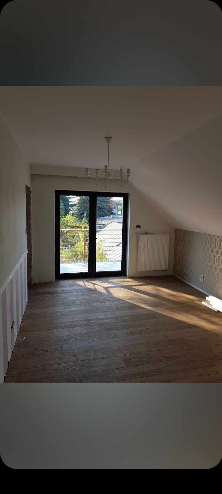 Pokój na poddaszu z drewnianą podłogą, balkonem z czarną ramą, grzejnikiem i wzorzystą tapetą na ścianie ze skosem.