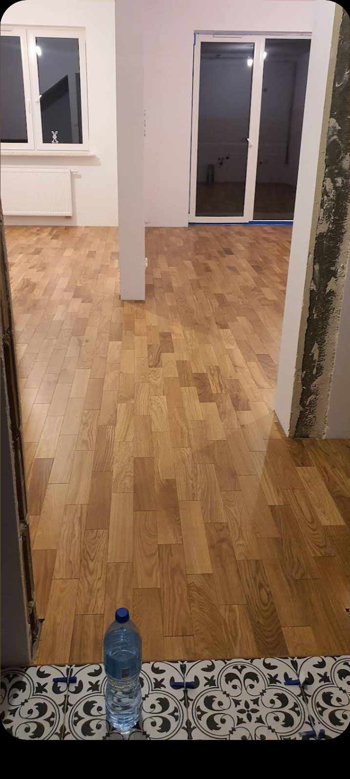 Ułożona drewniana podłoga w pustym pokoju, widoczna butelka wody w rogu, ściany przygotowane do malowania, okno i przeszklone drzwi balkonowe.