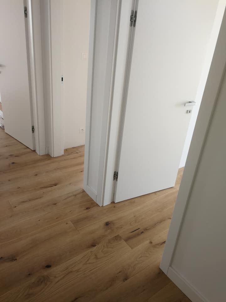 Jasna podłoga z paneli drewnopodobnych ułożona w jodełkę francuską, widok na otwarte białe drzwi wewnętrzne z srebrnymi klamkami i zawiasami.
