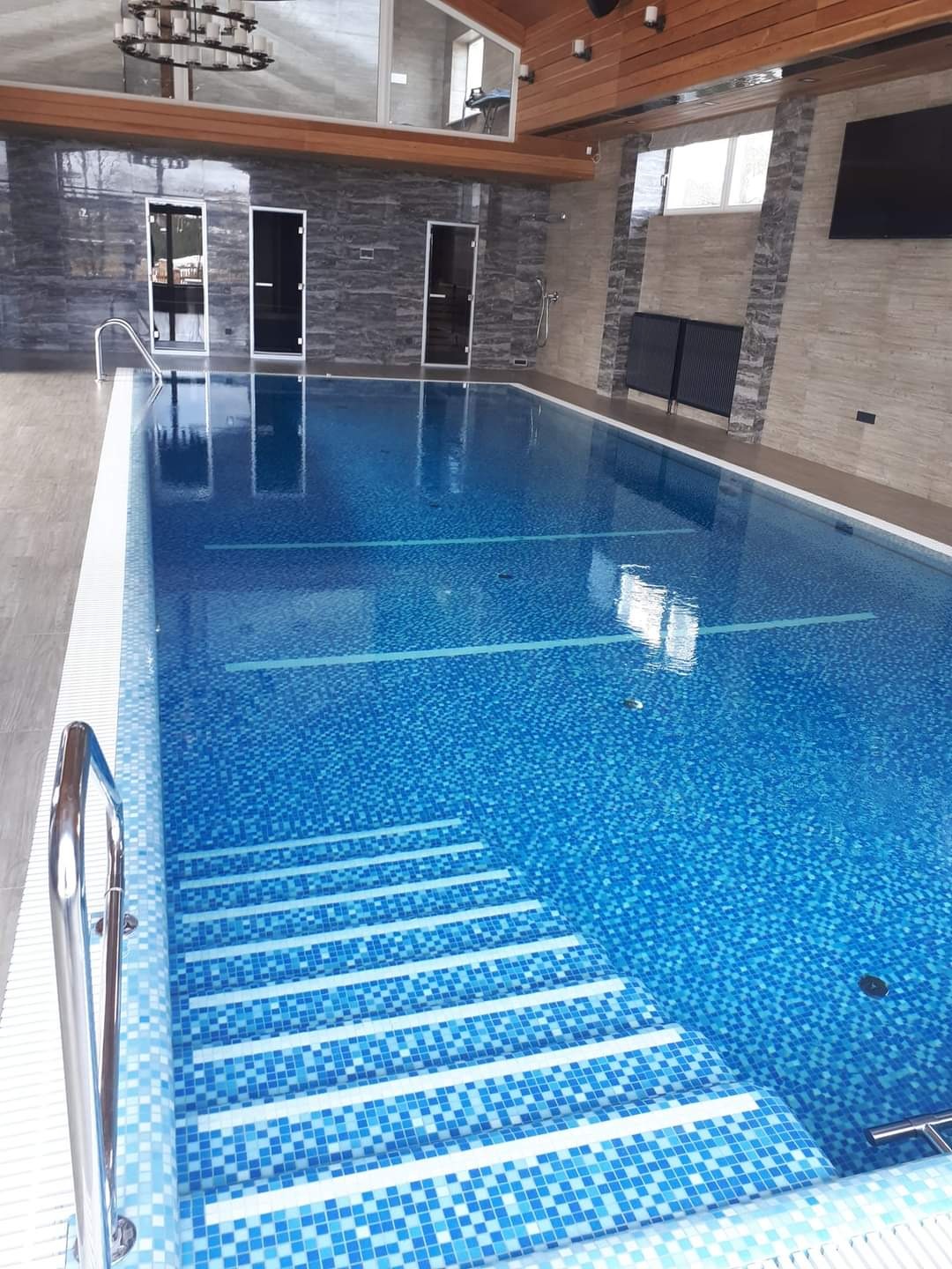 Błękitny basen z mozaiką, schodami i metalową poręczą, otoczony nowoczesnym wnętrzem z kamiennymi ścianami i drewnianym sufitem.