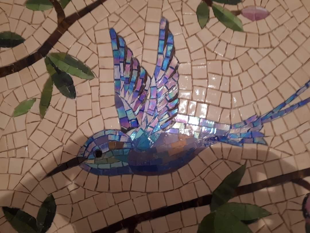 Mozaika ścienna przedstawiająca kolibra na gałęzi z liśćmi, wykonana z drobnych, nieregularnych płytek w odcieniach beżu, błękitu i fioletu.