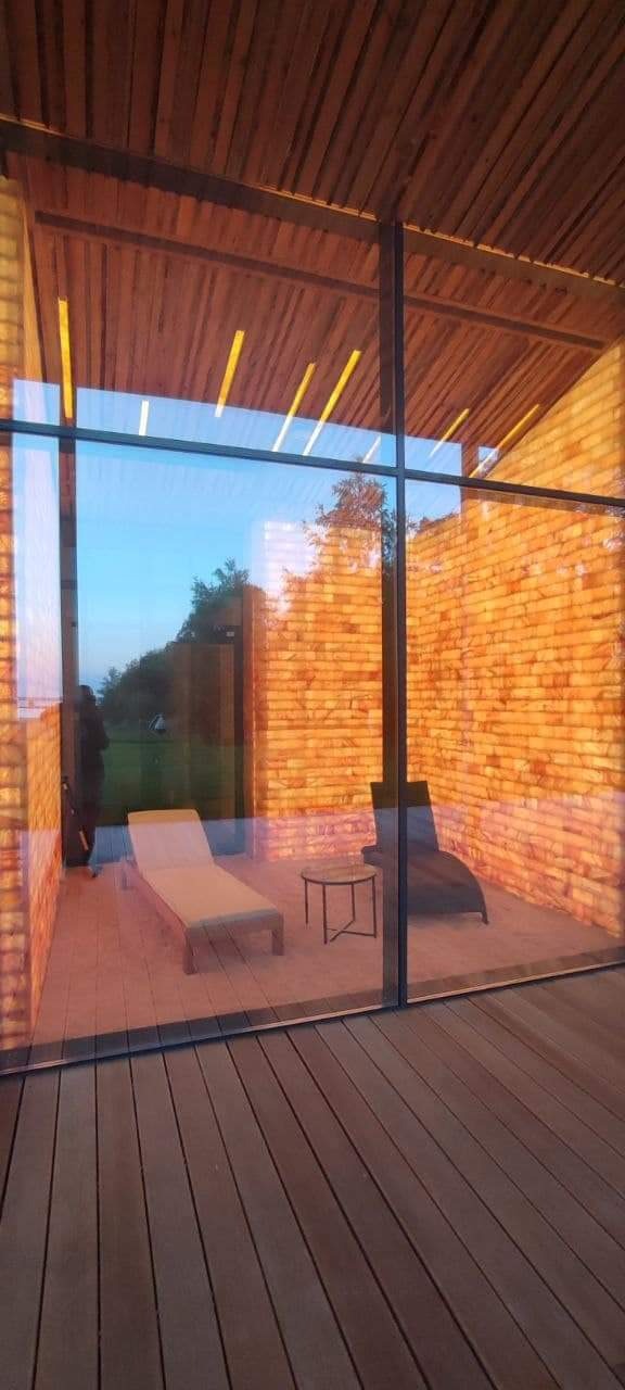 Nowoczesny taras z drewnianą podłogą i ceglaną ścianą, widoczny przez duże szklane okno, z leżakiem i stolikiem kawowym w środku, w tle zieleń.