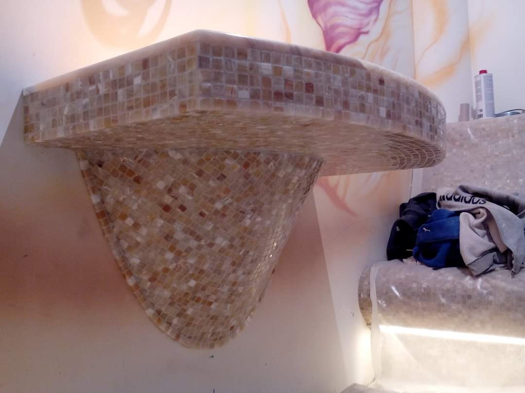 Narożna półka w łazience, wykończona mozaiką w odcieniach beżu i brązu, z widocznym fragmentem ściany z delikatnym motywem kwiatowym w tle.