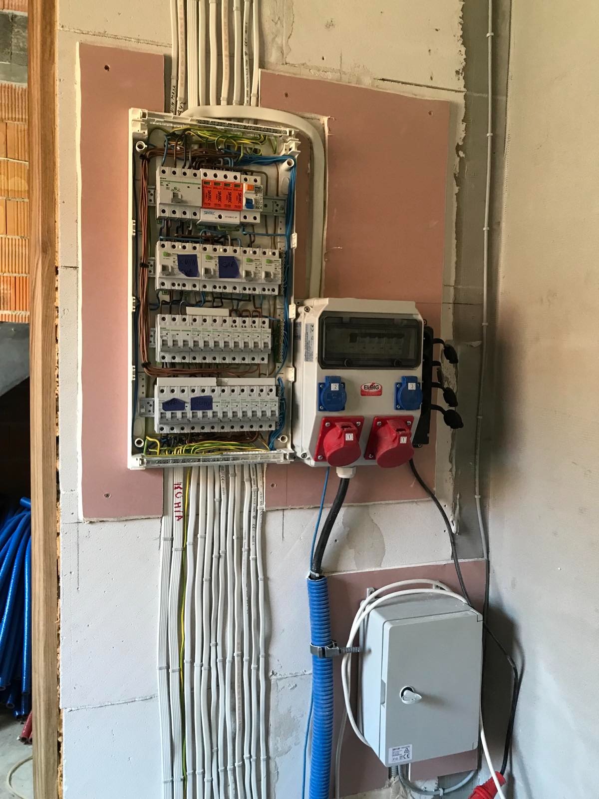 Rozdzielnica elektryczna z bezpiecznikami i gniazdami siłowymi zamontowana na ścianie pokrytej różową płytą kartonowo-gipsową, widoczne kable w peszlach i listwach.
