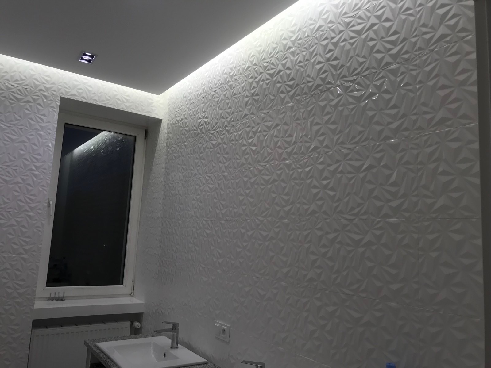 Nowoczesna łazienka z białymi, trójwymiarowymi płytkami na ścianach, podświetleniem LED i minimalistyczną umywalką.