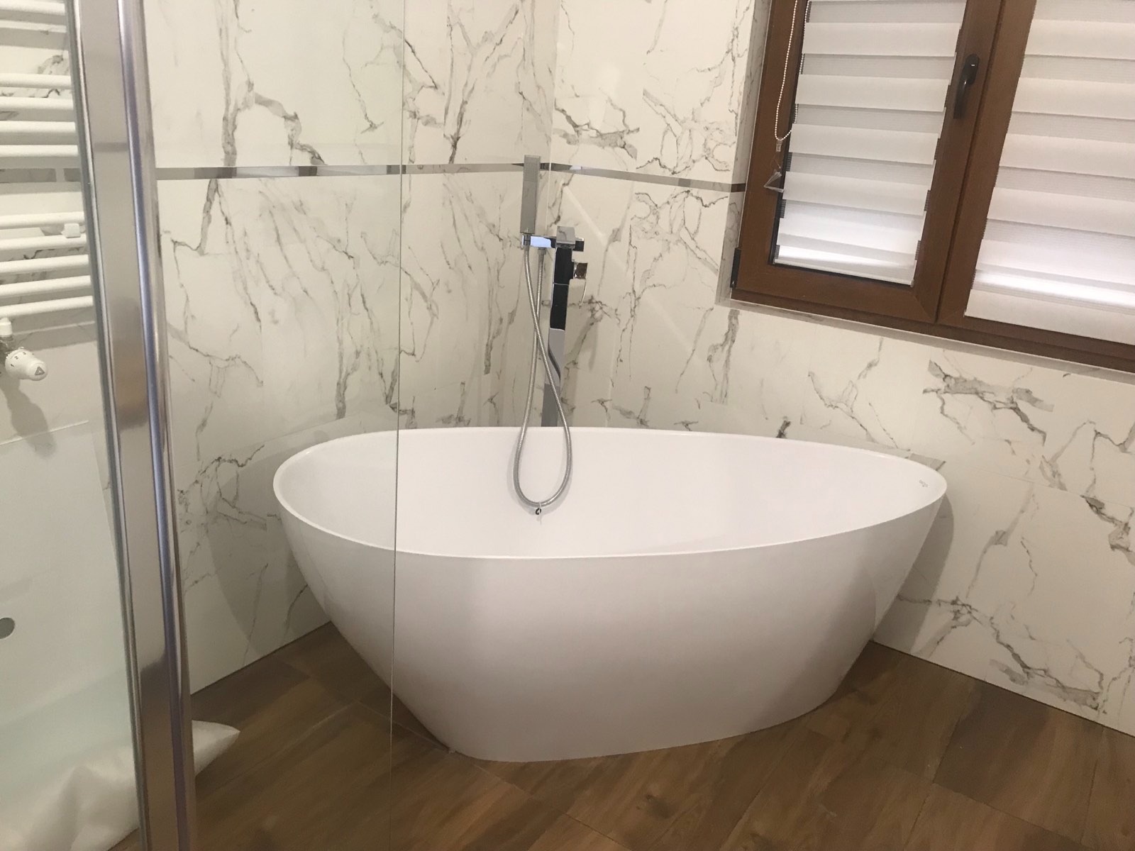 Wolnostojąca wanna owalna z chromowaną baterią na tle marmurowych płytek i drewnopodobnej podłogi, widok z boku, w tle okno z żaluzjami.