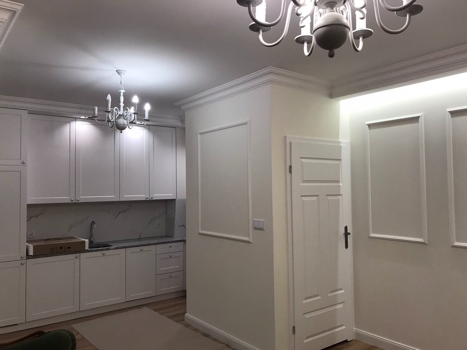 Jasne wnętrze po remoncie: kuchnia z białymi szafkami i marmurowym backsplash'em, ozdobne listwy przysufitowe, białe ściany z dekoracyjnymi ramkami i biała, klasyczna lampa sufitowa.