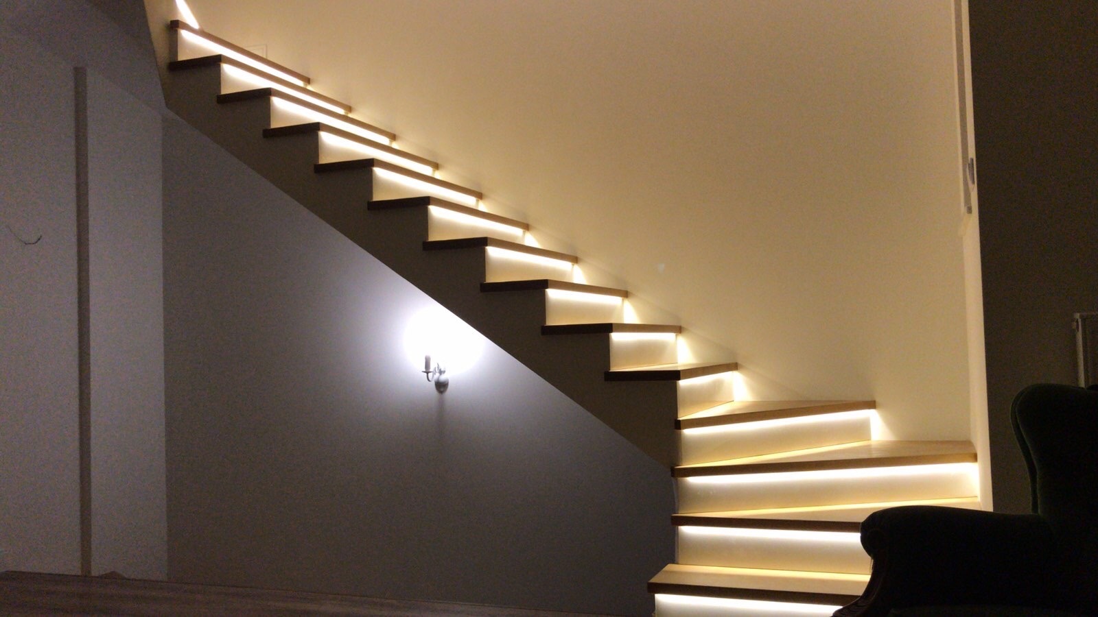 Nowoczesne schody z podświetleniem LED zamontowanym pod każdym stopniem, oświetlające ścianę i tworzące ciepłą, domową atmosferę. Na ścianie widoczny kinkiet.