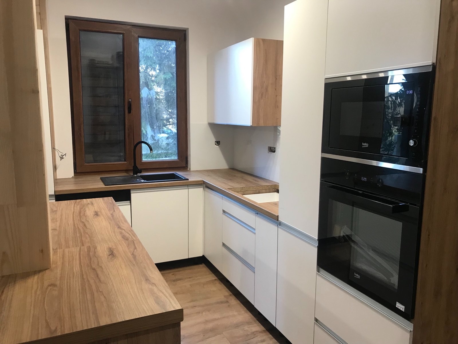 Nowoczesna kuchnia z białymi szafkami, drewnianym blatem i wbudowanym piekarnikiem oraz mikrofalówką marki Beko, z czarnym zlewem przy oknie.