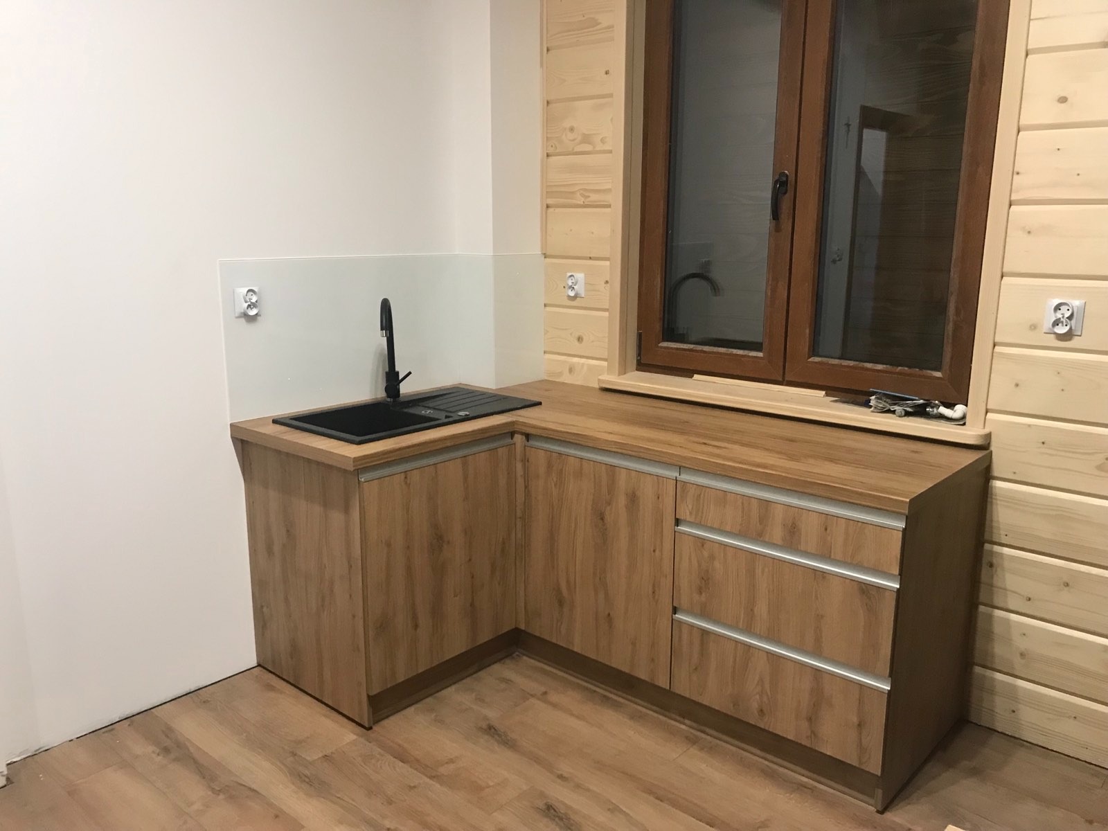 Nowoczesna zabudowa kuchenna w wykończeniu drewnopodobnym, z czarnym zlewem i armaturą, umieszczona pod oknem w drewnianej ramie, widok na fragment ściany z drewnianych paneli i białej ściany...