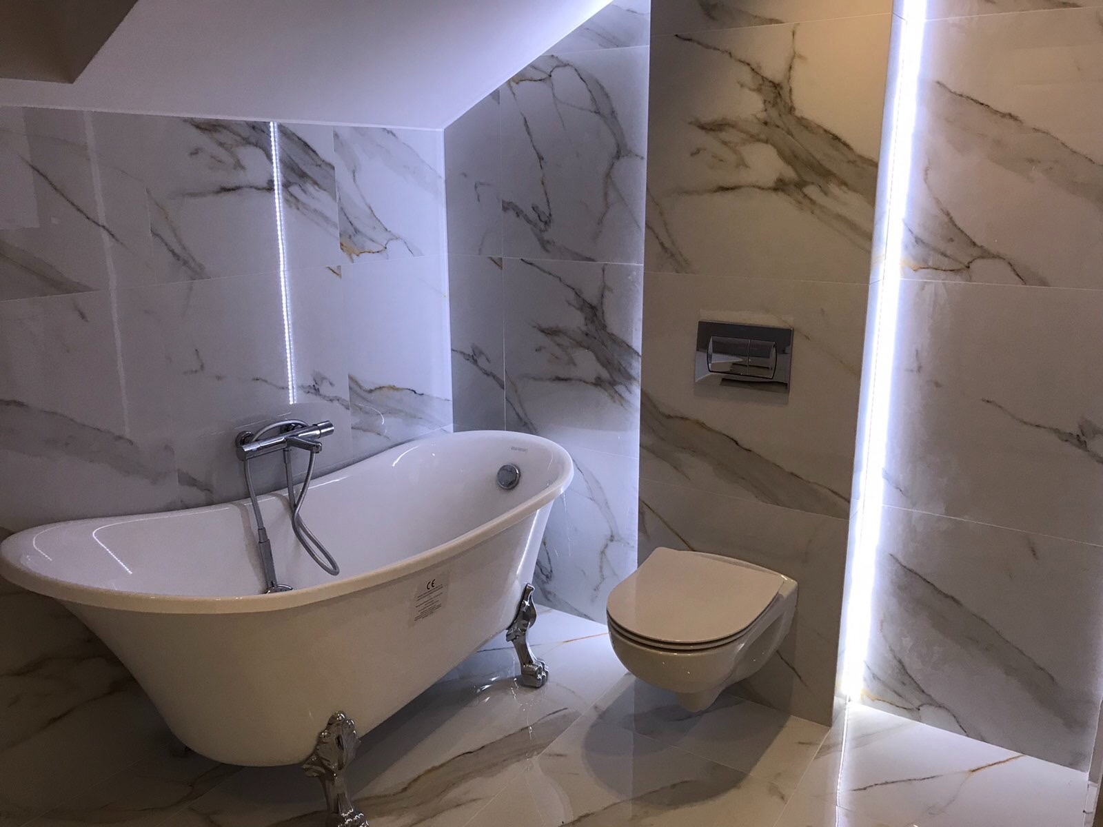 Elegancka łazienka w bieli i marmurze, z wolnostojącą wanną na ozdobnych nóżkach i podwieszaną toaletą, oświetlona pionowym paskiem ledowym.