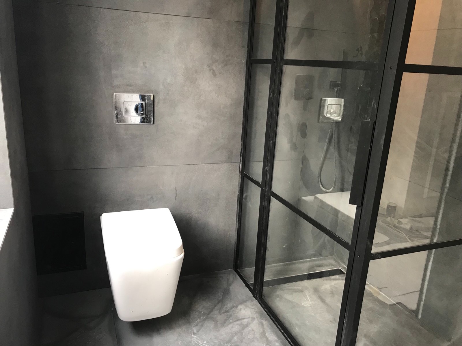 Nowoczesna łazienka w Warszawie z szarymi płytkami imitującymi beton, wisząca toaleta i kabina prysznicowa z czarnymi profilami.