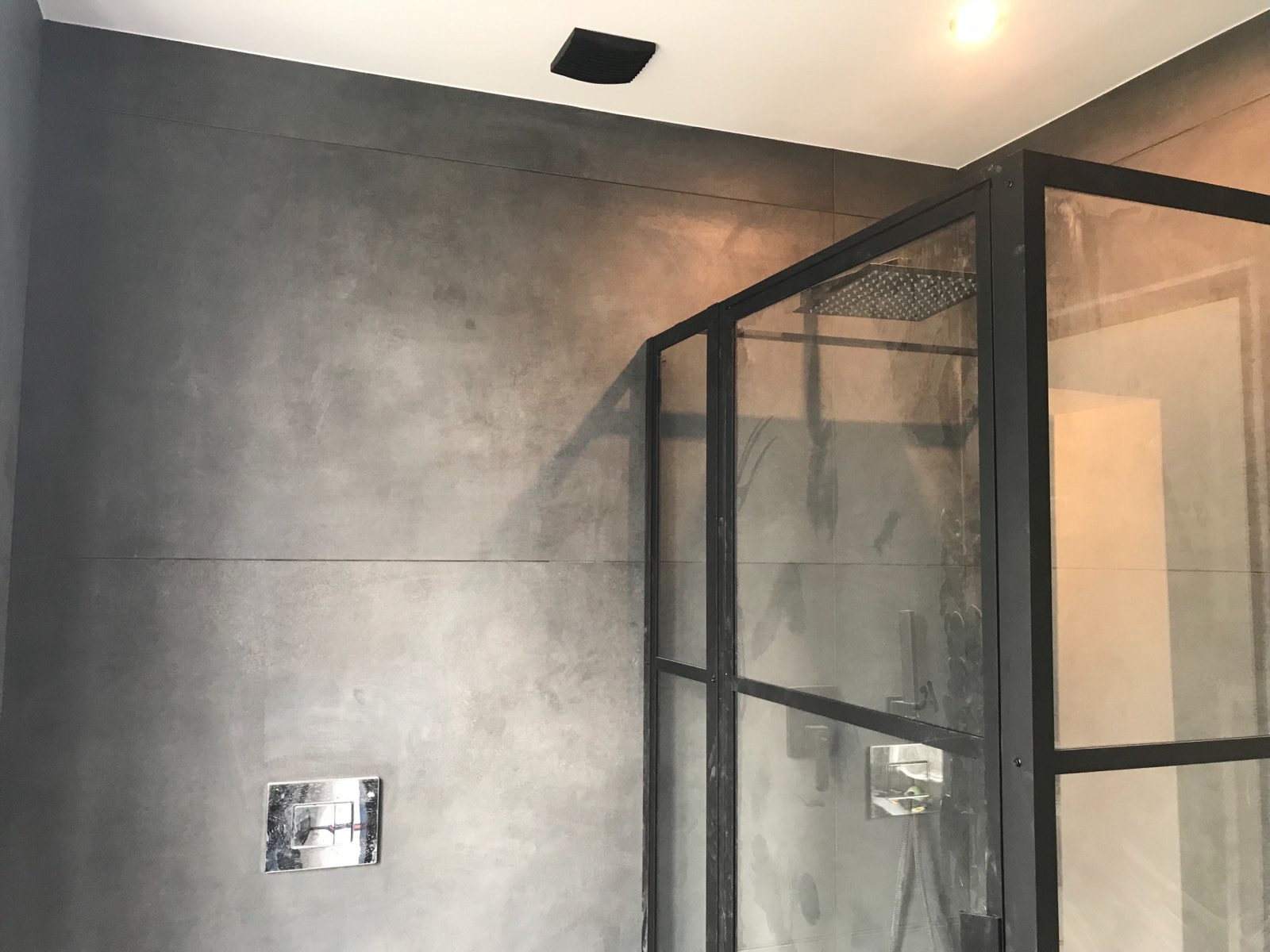 Nowoczesna łazienka z kabiną prysznicową typu walk-in z czarnymi profilami, szare płytki imitujące beton na ścianach, metalowy przycisk spłuczki.