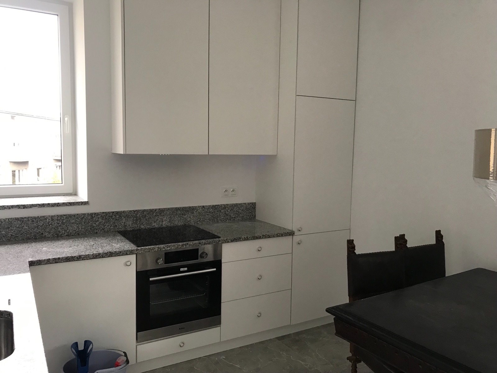 Nowoczesna biała kuchnia z granitowym blatem, czarną płytą indukcyjną i piekarnikiem Amica, widok na okno i fragment ciemnego, stylizowanego krzesła.
