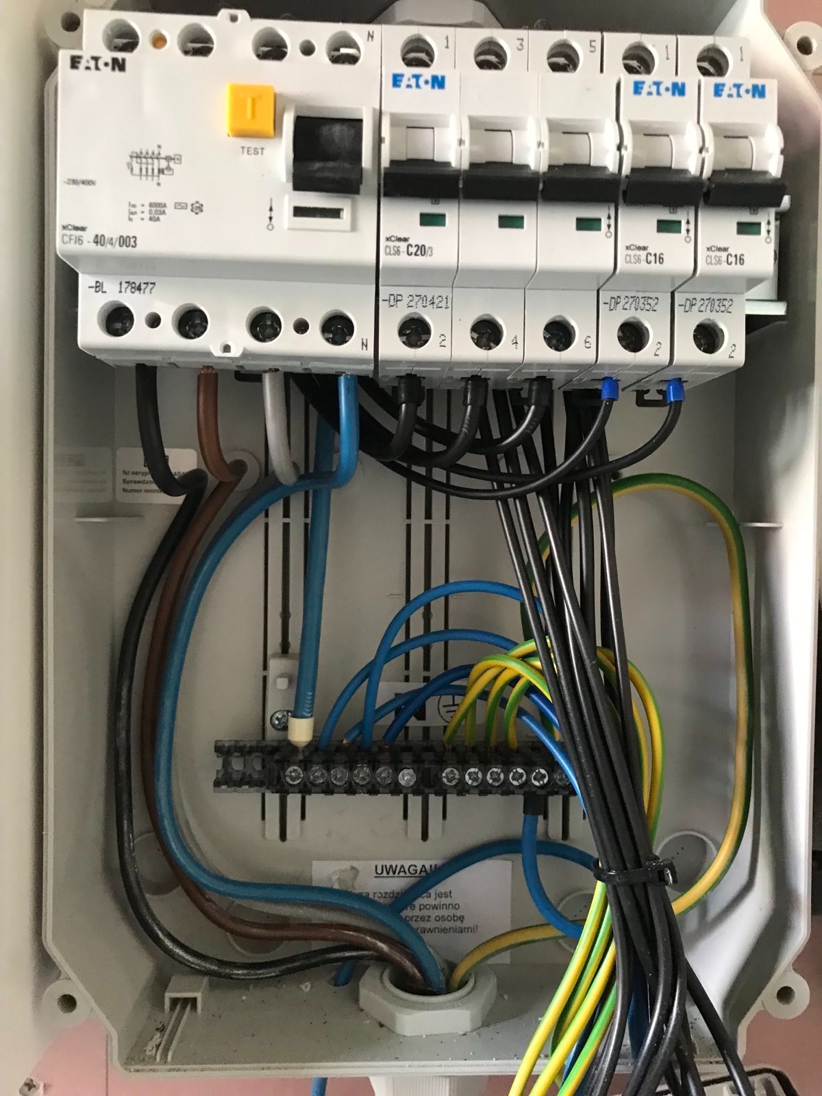 Rozdzielnica elektryczna z zainstalowanymi bezpiecznikami EATON, widoczne przewody w różnych kolorach podłączone do listwy zaciskowej, etykiety z numerami seryjnymi i ostrzeżeniem.