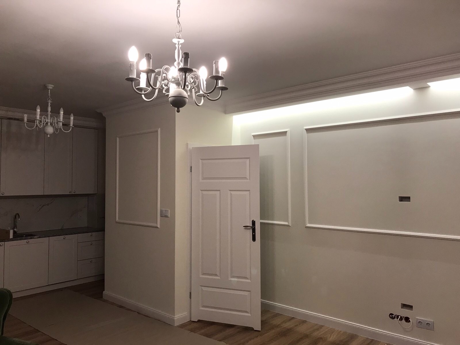 Wykończone wnętrze: kuchnia z białymi szafkami i marmurowym blatem, widoczny fragment salonu z białymi ścianami, sztukaterią, listwami przypodłogowymi, ozdobnymi ramami na ścianach, dwiema...