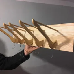 Ręcznie robiony wieszak z jasnego drewna z pięcioma haczykami, trzymany w dłoni na tle szarej ściany z fragmentem czerwonej zasłony po lewej stronie.