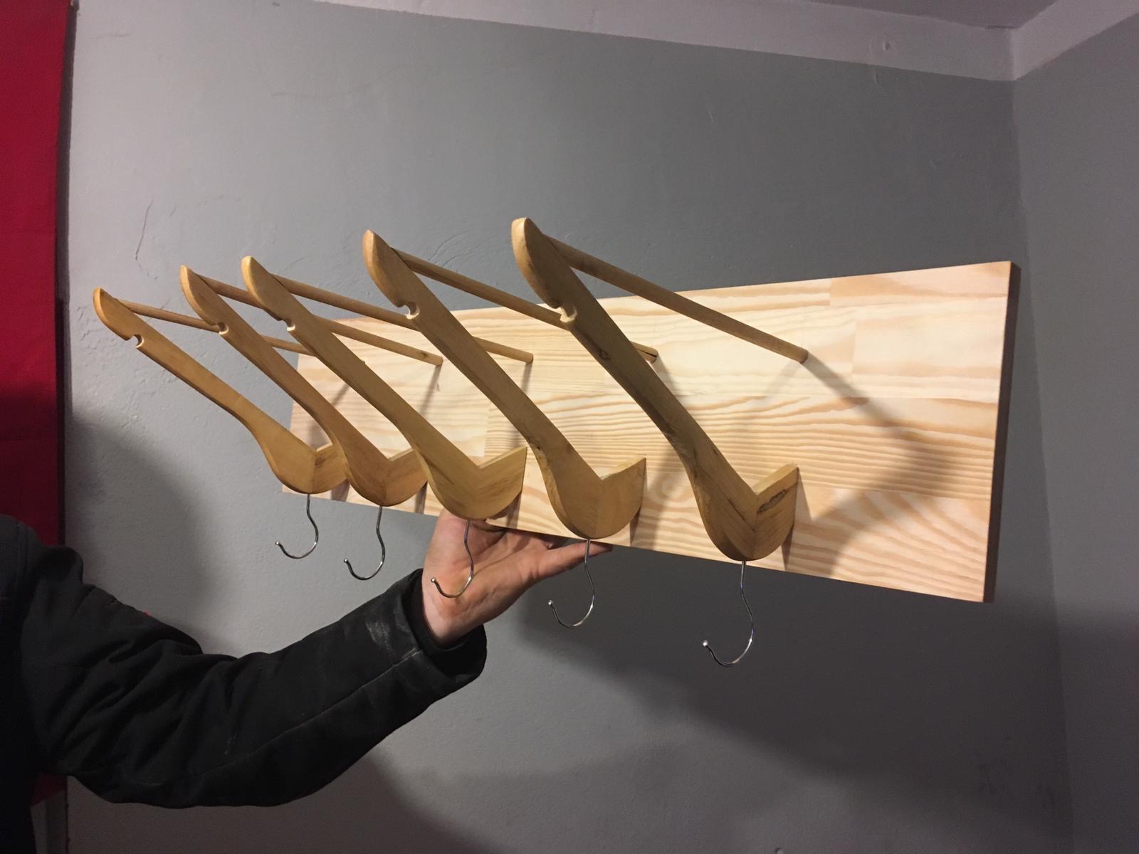 Ręcznie robiony wieszak z jasnego drewna z pięcioma haczykami, trzymany w dłoni na tle szarej ściany z fragmentem czerwonej zasłony po lewej stronie.