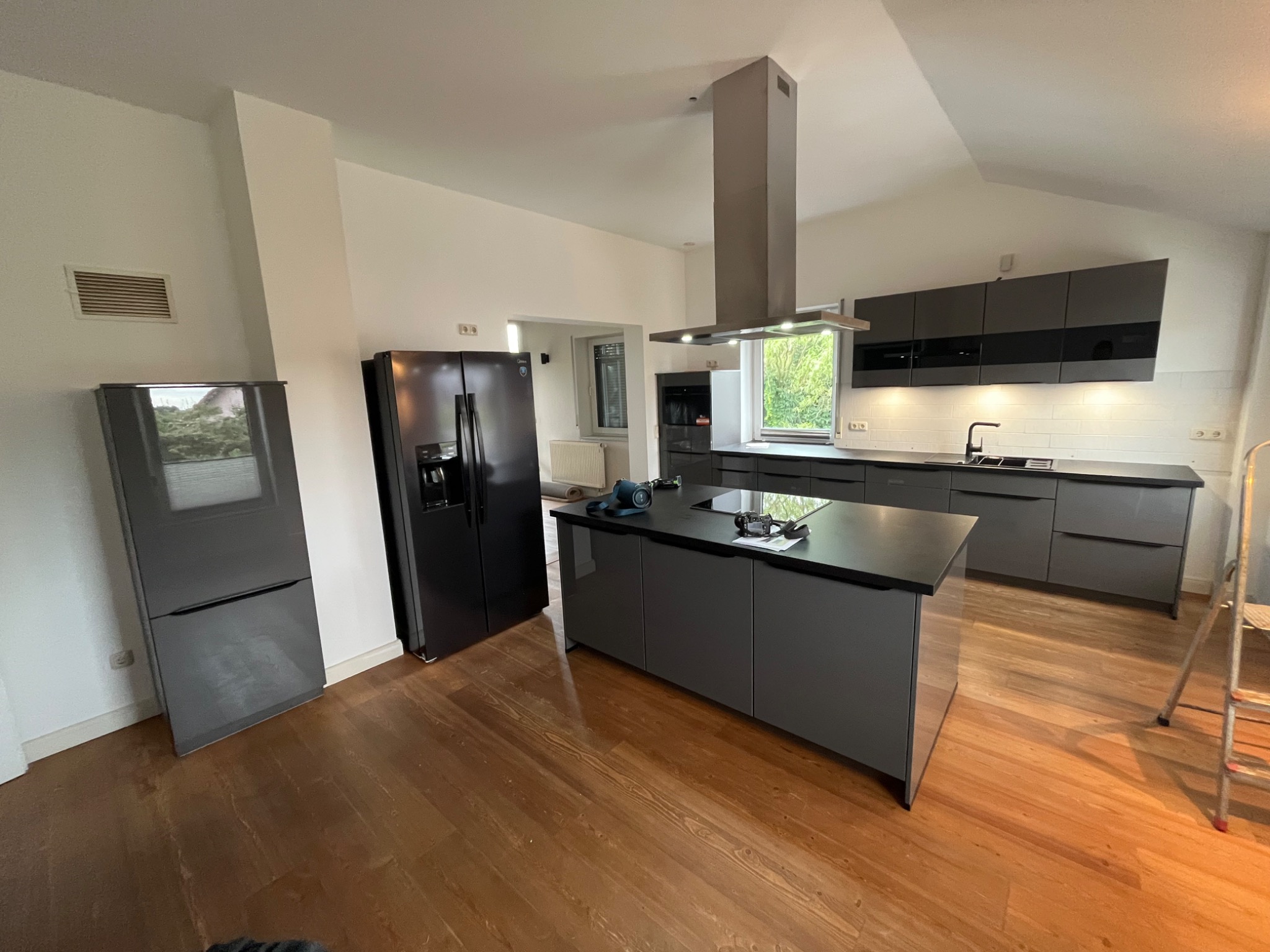 Nowoczesna kuchnia z szarymi szafkami, czarnym blatem i wyspą. Na blacie wyspy leżą narzędzia. W tle widoczny sprzęt AGD: lodówka i okap. Na podłodze drewniana podłoga.