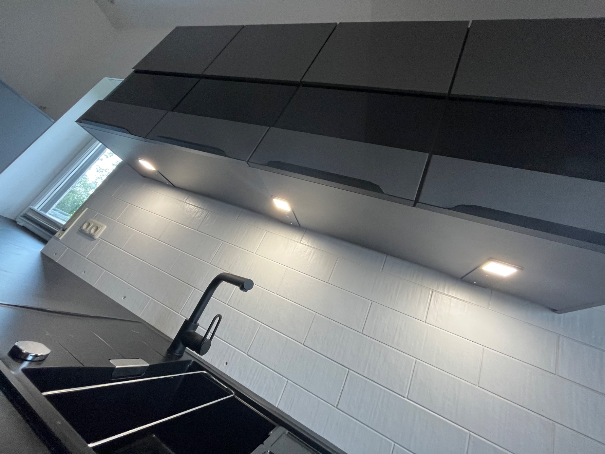 Nowoczesna kuchnia z czarnym zlewozmywakiem i baterią na tle białych, prostokątnych płytek oraz górnych szafek w odcieniach szarości z oświetleniem punktowym.