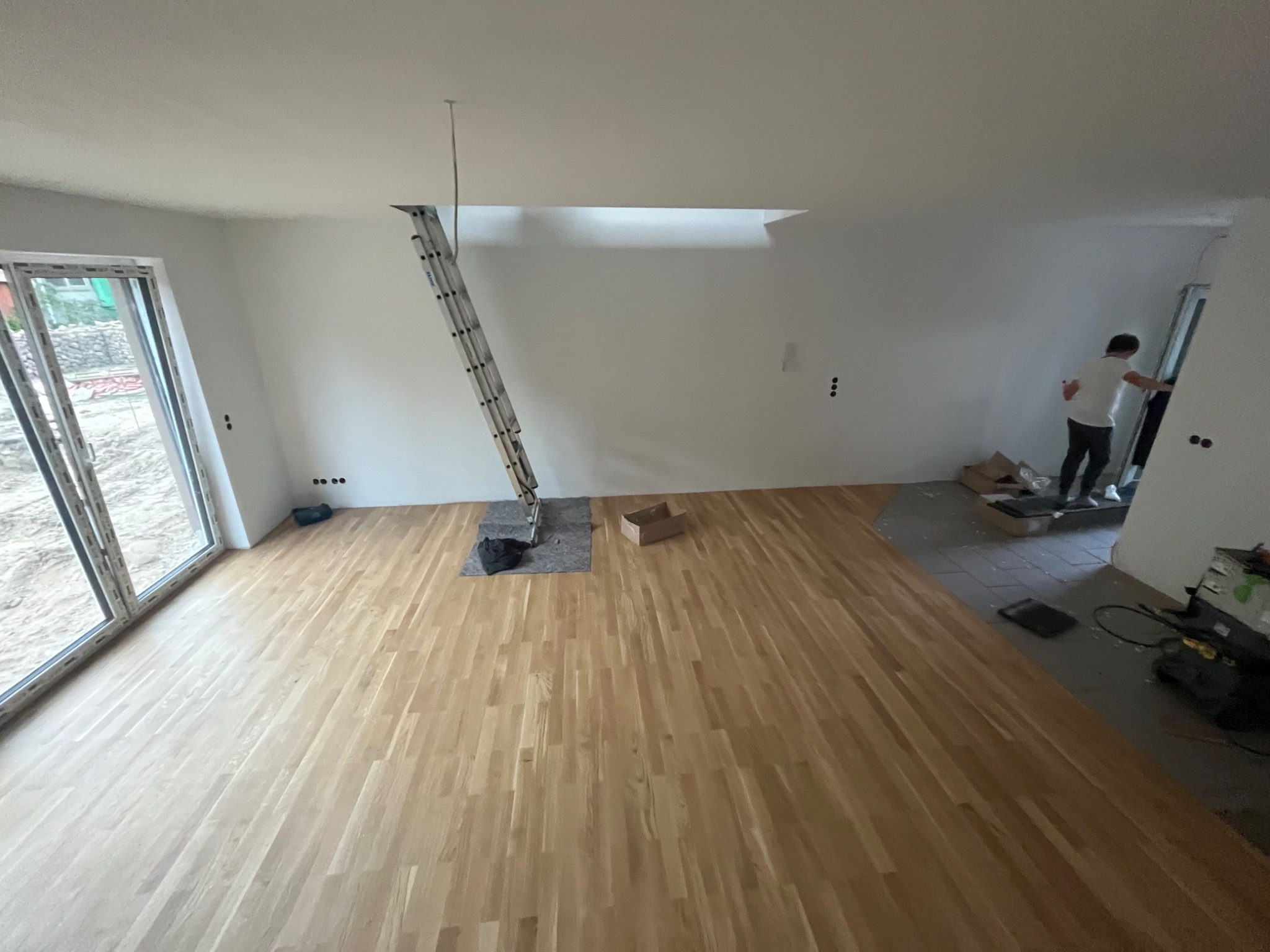 Nowo położona, jasna drewniana podłoga w przestronnym pokoju z dużym oknem; drabina oparta o ścianę sugeruje prace wykończeniowe przy suficie lub instalacji oświetlenia.