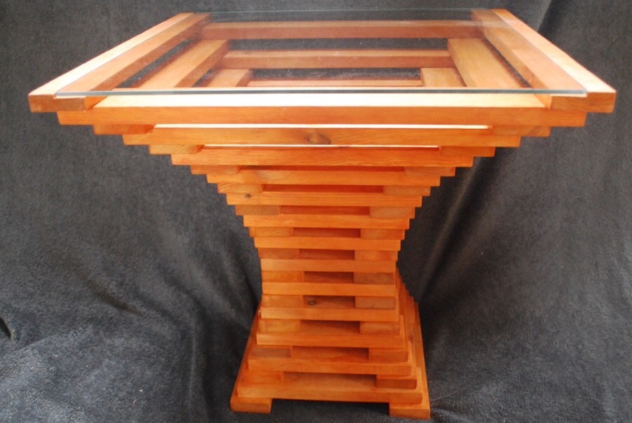 Nietypowy stolik kawowy z drewnianych listewek ułożonych schodkowo i szklanym blatem, na ciemnoszarym tle.