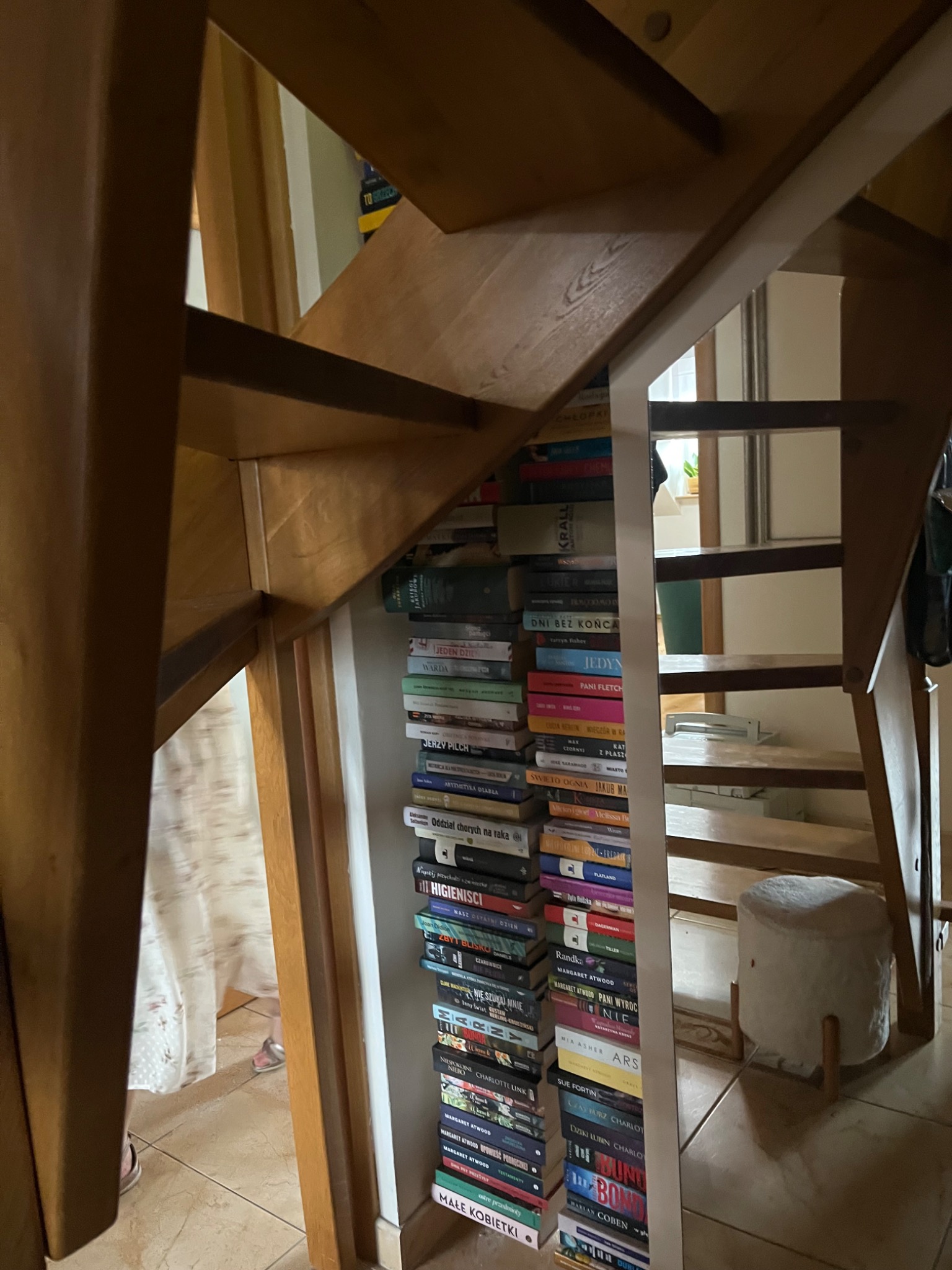 Drewniane schody z wbudowanym regałem na książki, wypełnionym po brzegi różnorodnymi tytułami, widoczne częściowo przez szczeliny konstrukcji schodów.