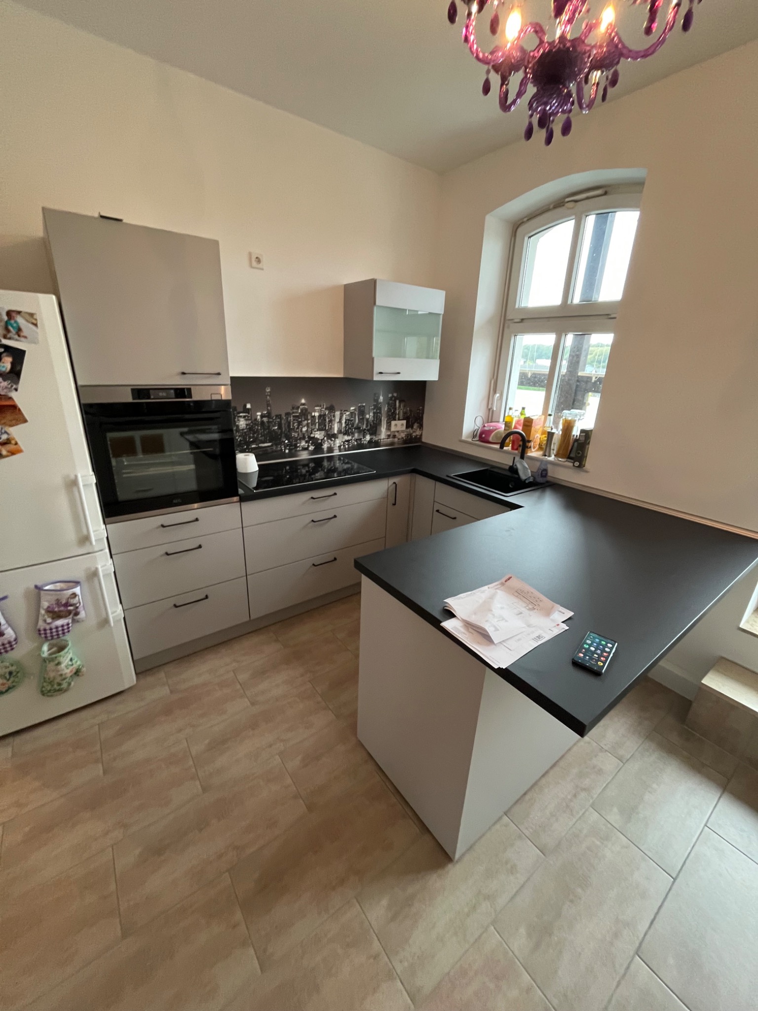 Jasna kuchnia z szarymi szafkami, czarnym blatem, czarnym panelem ściennym z motywem miasta, widok na okno i fioletowy żyrandol, na blacie leżą dokumenty i telefon.