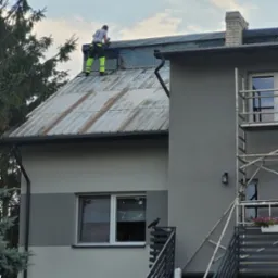 Pracownik w odblaskowych spodniach na dachu domu, wykonuje prace dekarskie, w tle komin, na schodach kruk.