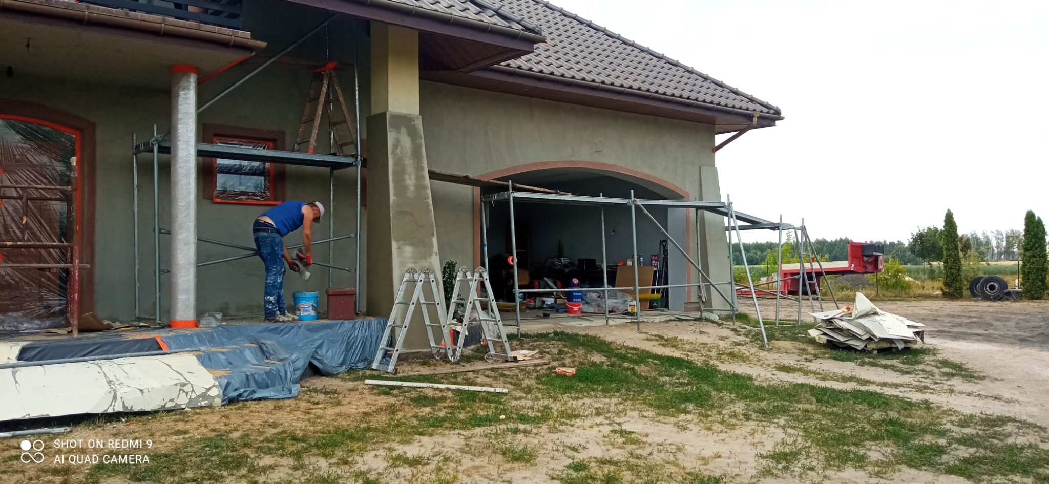 Pracownik w niebieskiej koszulce i dżinsach szpachluje ścianę domu ze stojącego rusztowania, widoczna wiadro z zaprawą, zabezpieczone okna folią i bałagan na posesji.