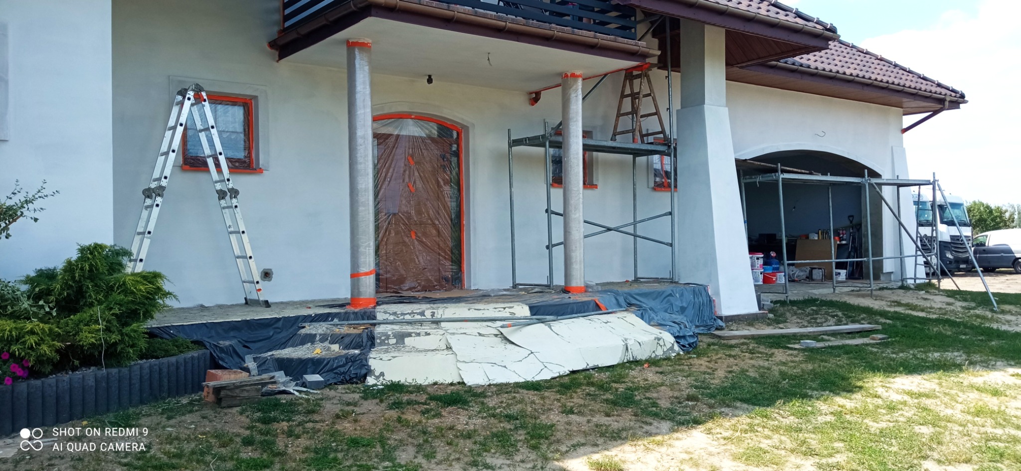 Dom jednorodzinny w trakcie prac elewacyjnych: okna i drzwi zabezpieczone folią, kolumny owinięte, widoczne rusztowania i drabina oparta o świeżo otynkowaną ścianę.
