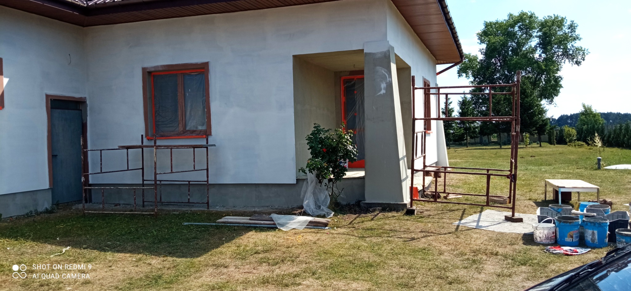Dom jednorodzinny w trakcie tynkowania elewacji, z widocznymi rusztowaniami, zabezpieczonymi oknami i przygotowanymi pojemnikami z zaprawą.
