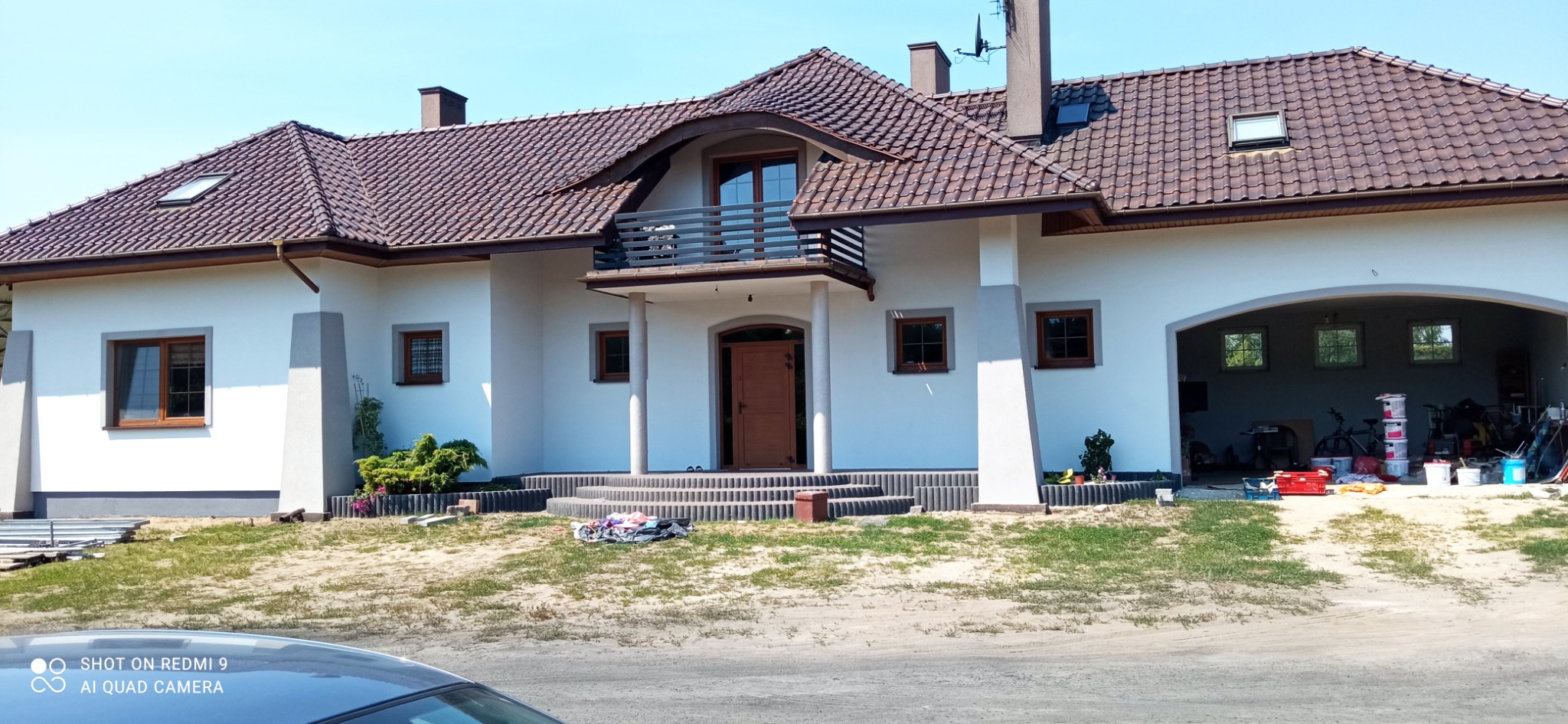 Biały dom jednorodzinny z brązowym dachem i stolarką okienną, widoczne świeżo pomalowane ściany i bałagan w otwartym garażu.