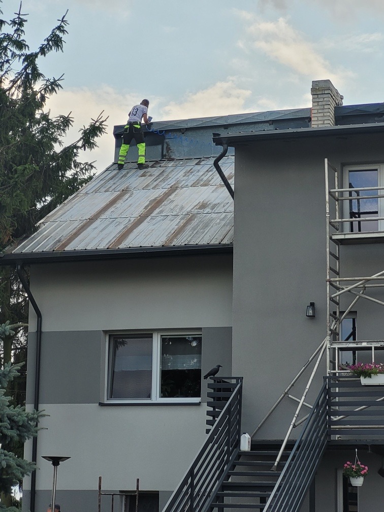 Pracownik w odblaskowych spodniach na dachu domu, wykonuje prace dekarskie, w tle komin, na schodach kruk.