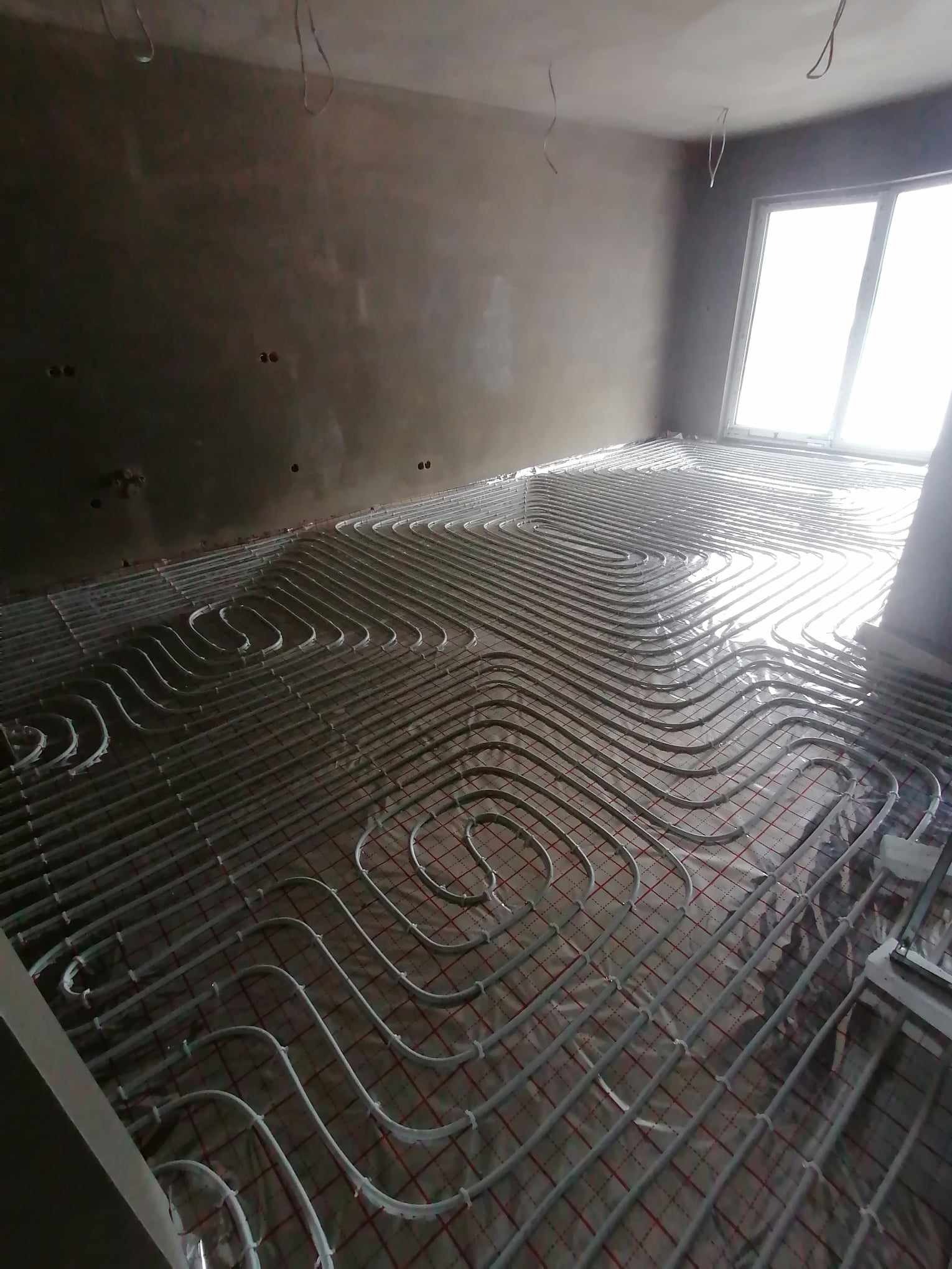 Szare rury ogrzewania podłogowego ułożone na folii izolacyjnej w surowym wnętrzu z widocznymi przewodami elektrycznymi w suficie i oknem w tle.