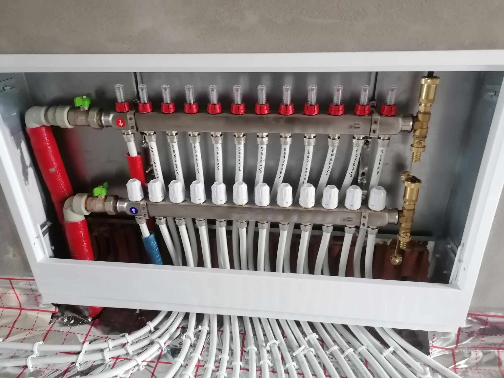Rozdzielacz ogrzewania podłogowego z zaworami i rurami, zamontowany w białej skrzynce, widoczne oznaczenia pomieszczeń na rurach.