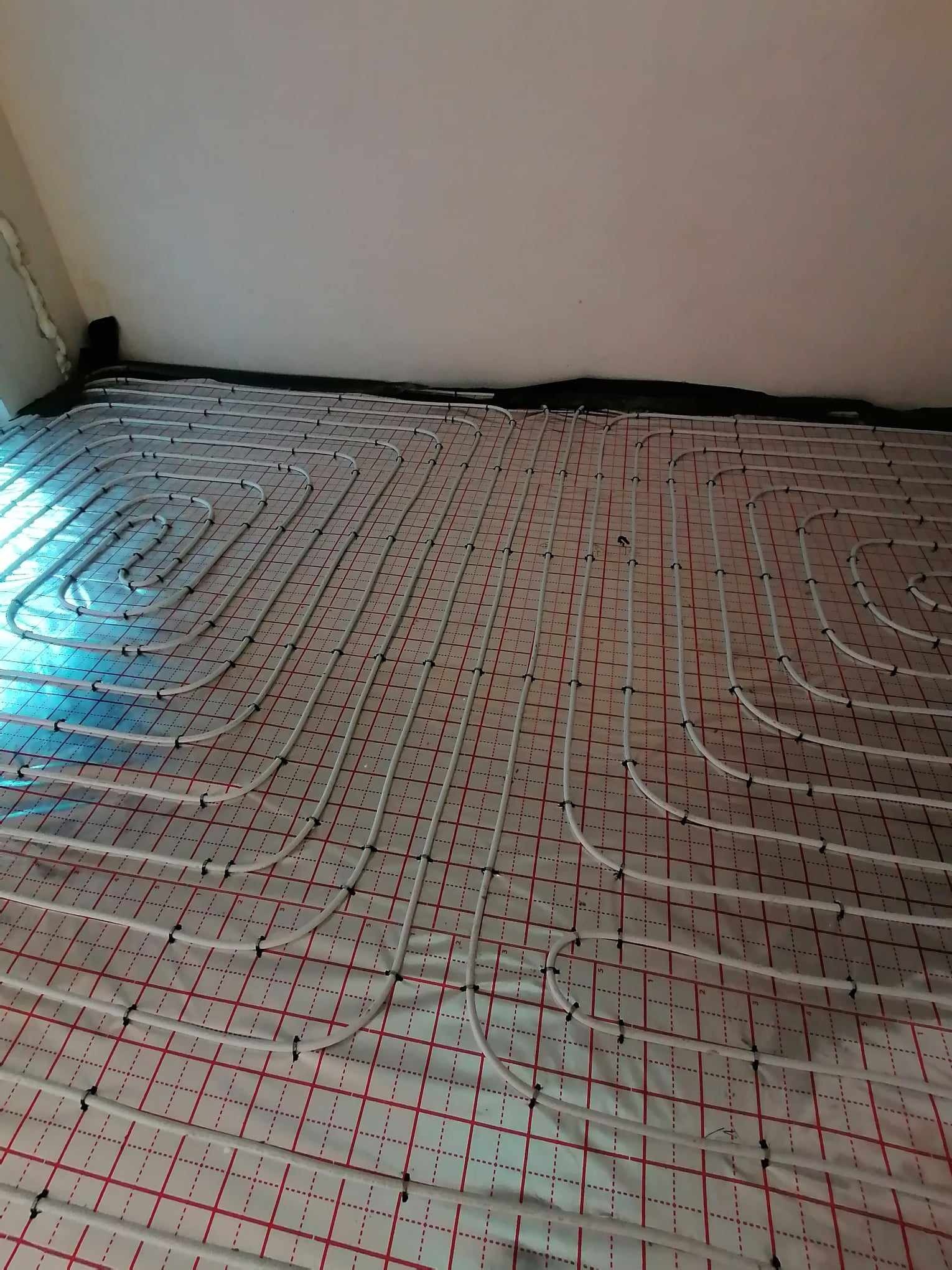 Białe rurki ogrzewania podłogowego, zamocowane czarnymi klipsami na folii z czerwoną siatką, rozłożone na przygotowanym podłożu w pomieszczeniu.