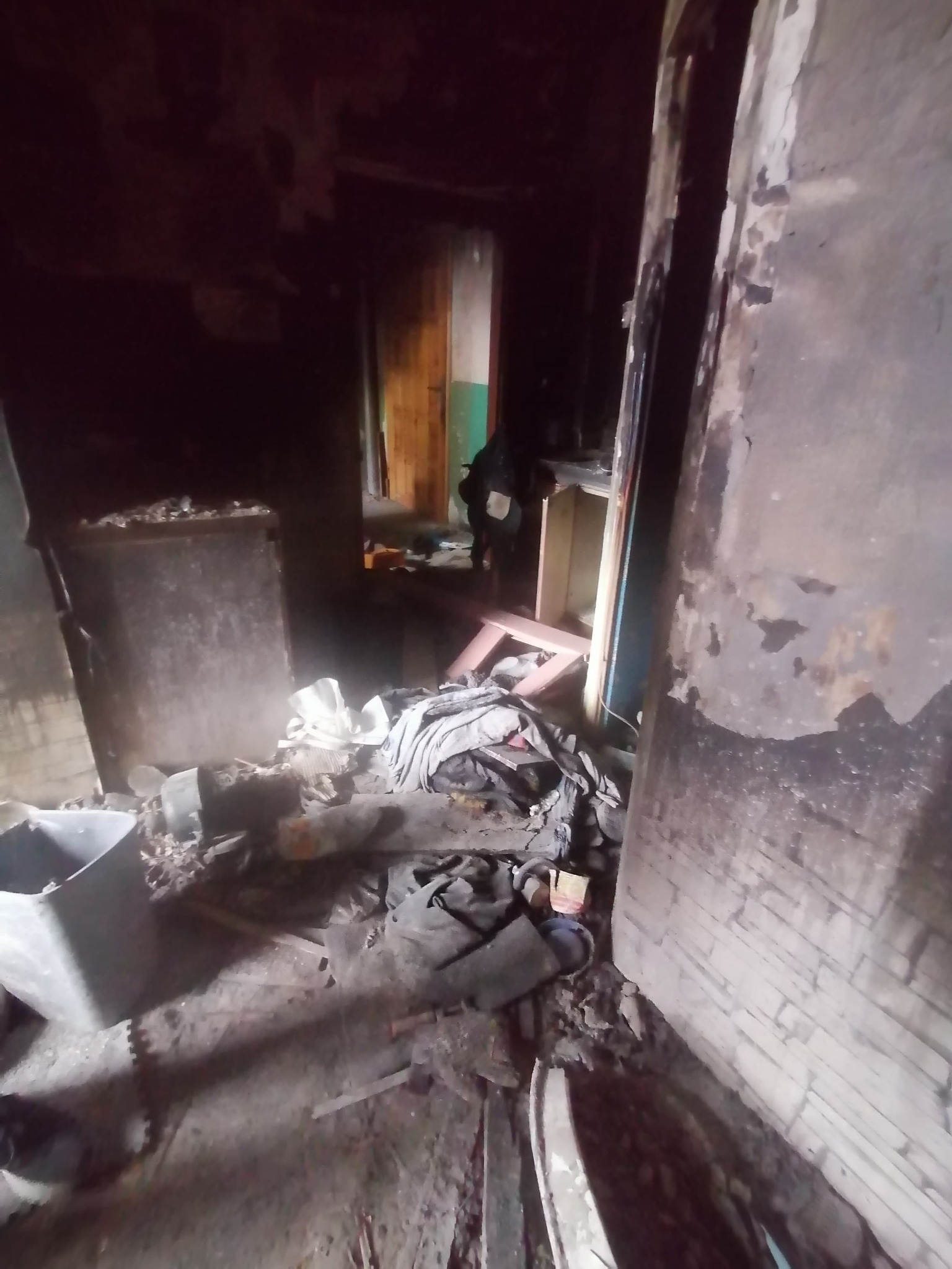 Wnętrze zniszczonego pomieszczenia po pożarze, widoczne zwęglone ściany, gruz, pozostałości mebli i odpadów, słabe oświetlenie.
