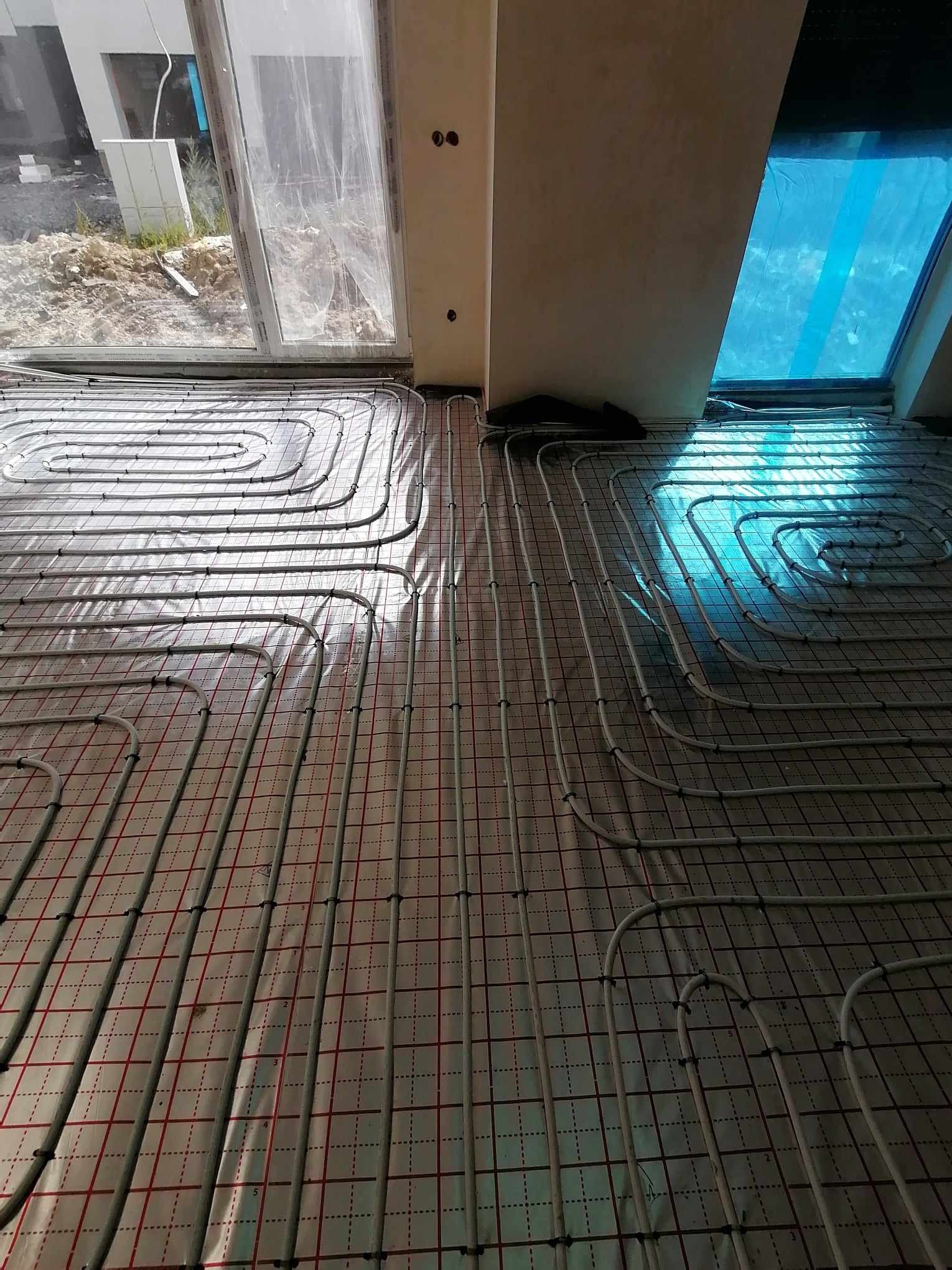 Białe rury ogrzewania podłogowego ułożone spiralnie na folii z czerwoną siatką, widoczne okna i drzwi balkonowe w tle.