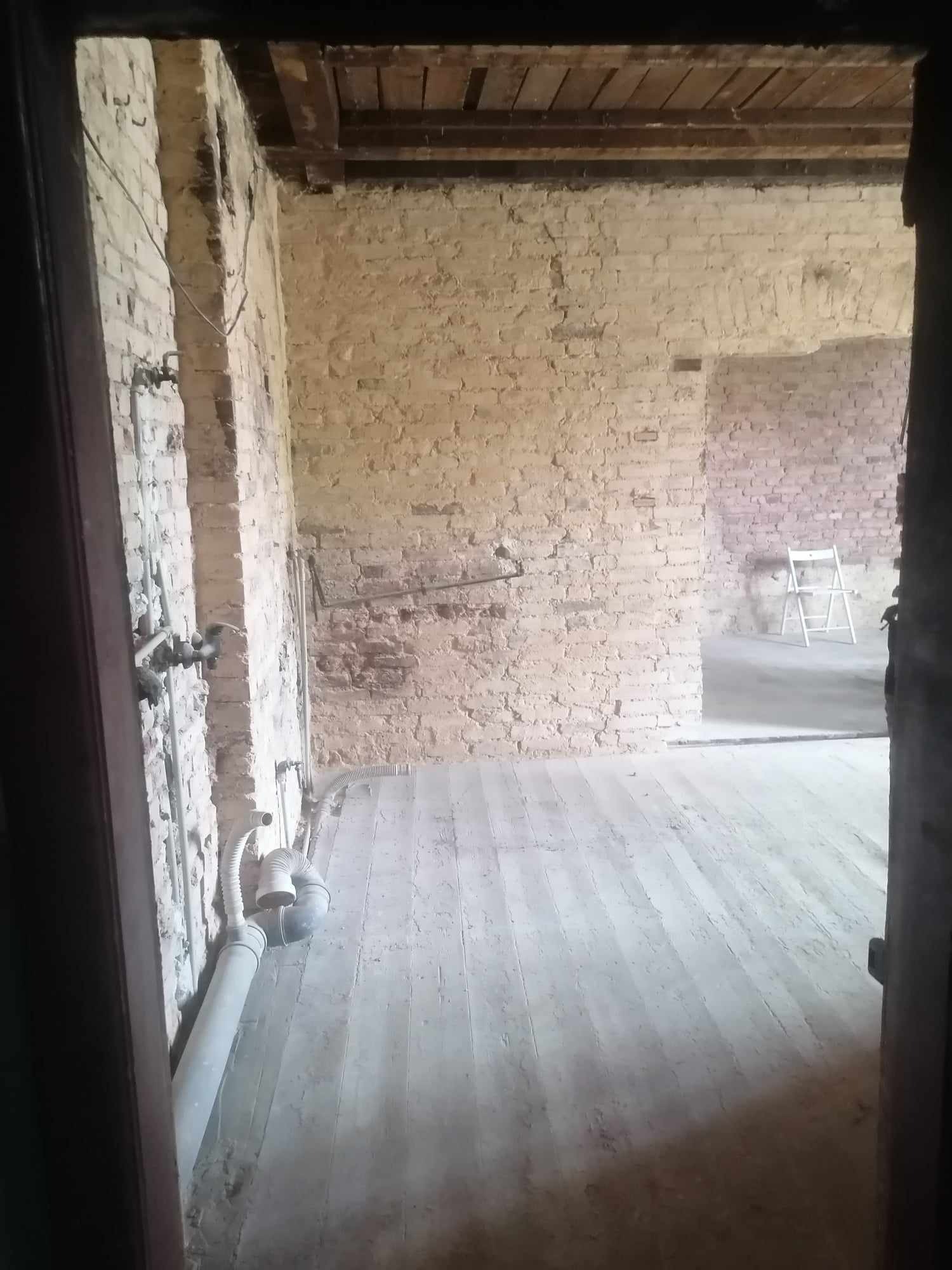 Wnętrze remontowanego pomieszczenia z odsłoniętą cegłą, widoczne instalacje hydrauliczne i drewniana podłoga, białe krzesło w oddali.