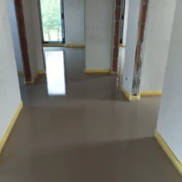 Świeżo wylana, mokra wylewka anhydrytowa w jasnym pomieszczeniu z widocznymi oknami i fragmentami ścian zabezpieczonych taśmą malarską.