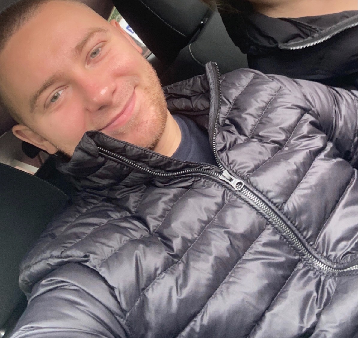 Uśmiechnięty mężczyzna w czarnej, błyszczącej kurtce puchowej siedzący na fotelu samochodowym, z widocznym fragmentem drugiego pasażera w tle.