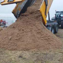 Żółta wywrotka w trakcie wysypywania dużej ilości piasku na pole, w tle widoczny traktor i tunel foliowy, pochmurne niebo.