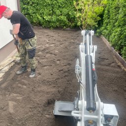 Ogrody - Przygotowanie terenu pod trawnik: pracownik wyrównuje ziemię łopatą obok mini koparki, w tle zielone tuje i ściana budynku.
