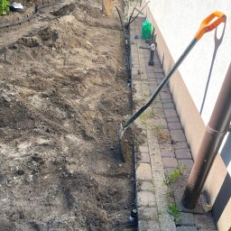 Ogrody - Wykop pod instalację nawadniającą wzdłuż ściany, z widocznymi rurami i zraszaczami. Na pierwszym planie łopata z pomarańczową rączką. Widoczna zieleń i elementy budynku.