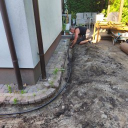 Ogrody - Mężczyzna instaluje system nawadniania ogrodu, układając czarną rurę w wykopanym rowie wzdłuż ściany domu z rynnami. Widoczne narzędzia i częściowo wyposażony taras.