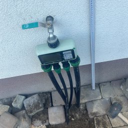 Ogrody - Podłączenie systemu nawadniania: kran z elektronicznym sterownikiem i czterema wężami, obok leżą kostki brukowe i miara zwinięta na ścianie.