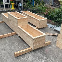 Ogrody - Ręcznie robione, surowe drewniane skrzynie na rośliny ogrodowe, ustawione na betonowym podłożu, z widocznymi elementami konstrukcyjnymi i zielenią w tle.