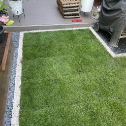 Ogrody - Świeża trawa z rolki otoczona kamienną obwódką, przylegająca do drewnianego tarasu i grilla. Fragment rabaty z roślinami w tle. Widok z góry, słoneczny dzień.