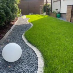 Falista ścieżka z szarego żwiru, obrzeża z kostki, trawnik z soczystą zielenią, oświetlony kulami, w tle drewniane ogrodzenie i pergola.